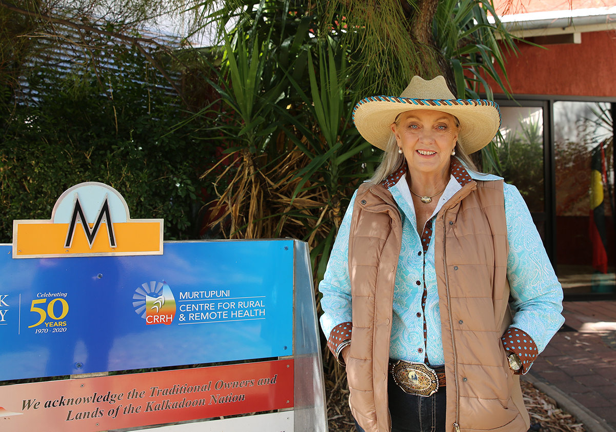 Sabina wearing a wide brimmed had, shirt, and belt with large decorative buckle, standing next to the Murtupuni Centre for Rural and Remote Health sign.