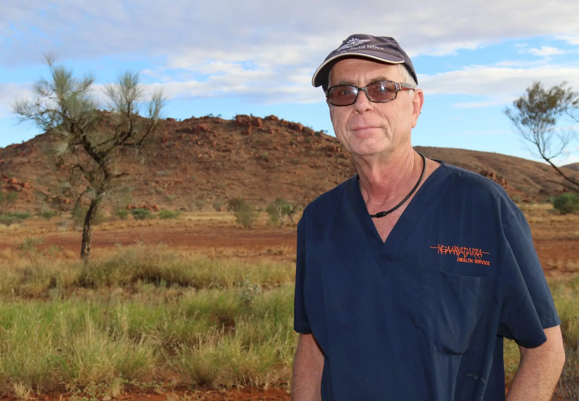 Man in a blue scrub shirt that says Ngaanyatjarra health service and an RFDS cap on, with remote landscape in background.