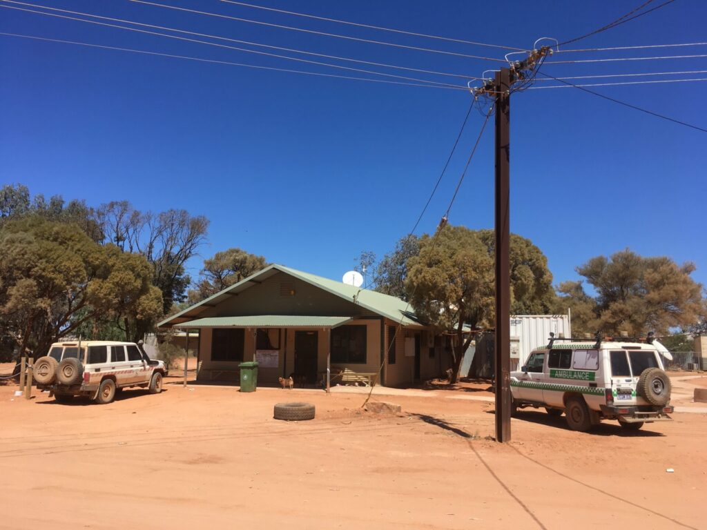 A remote clinic building surrounded by red dirt and gum tress with two 4WD ambulances parked outside.