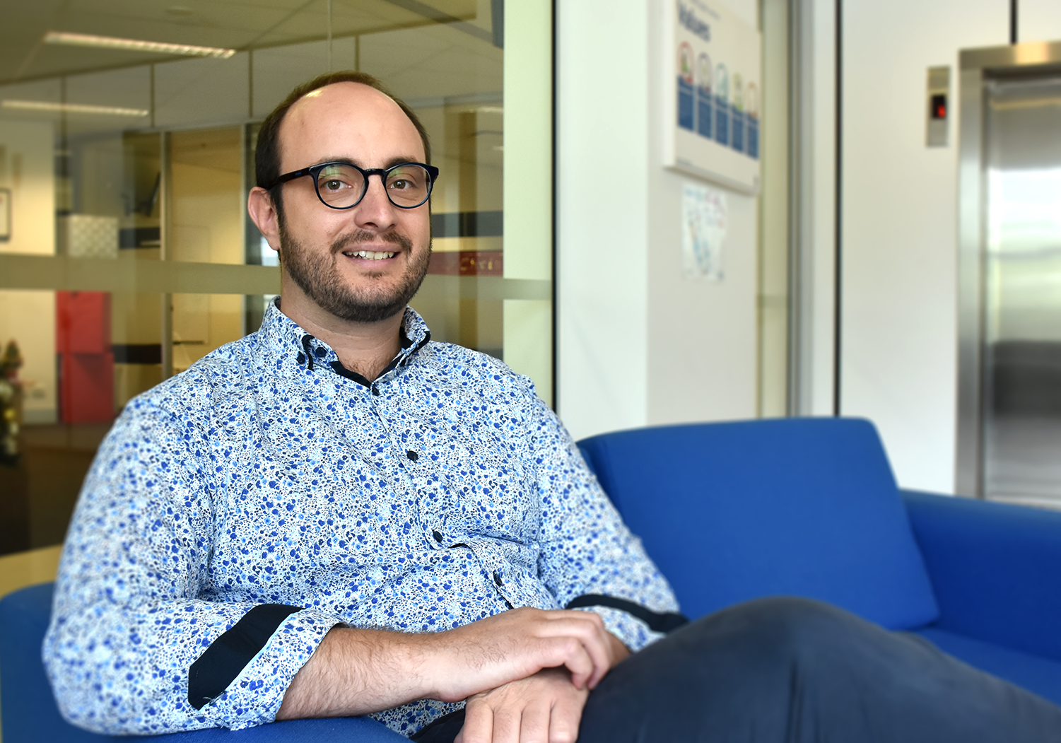 Keppel in a blue shirt, sat on a blue sofa, smiling.
