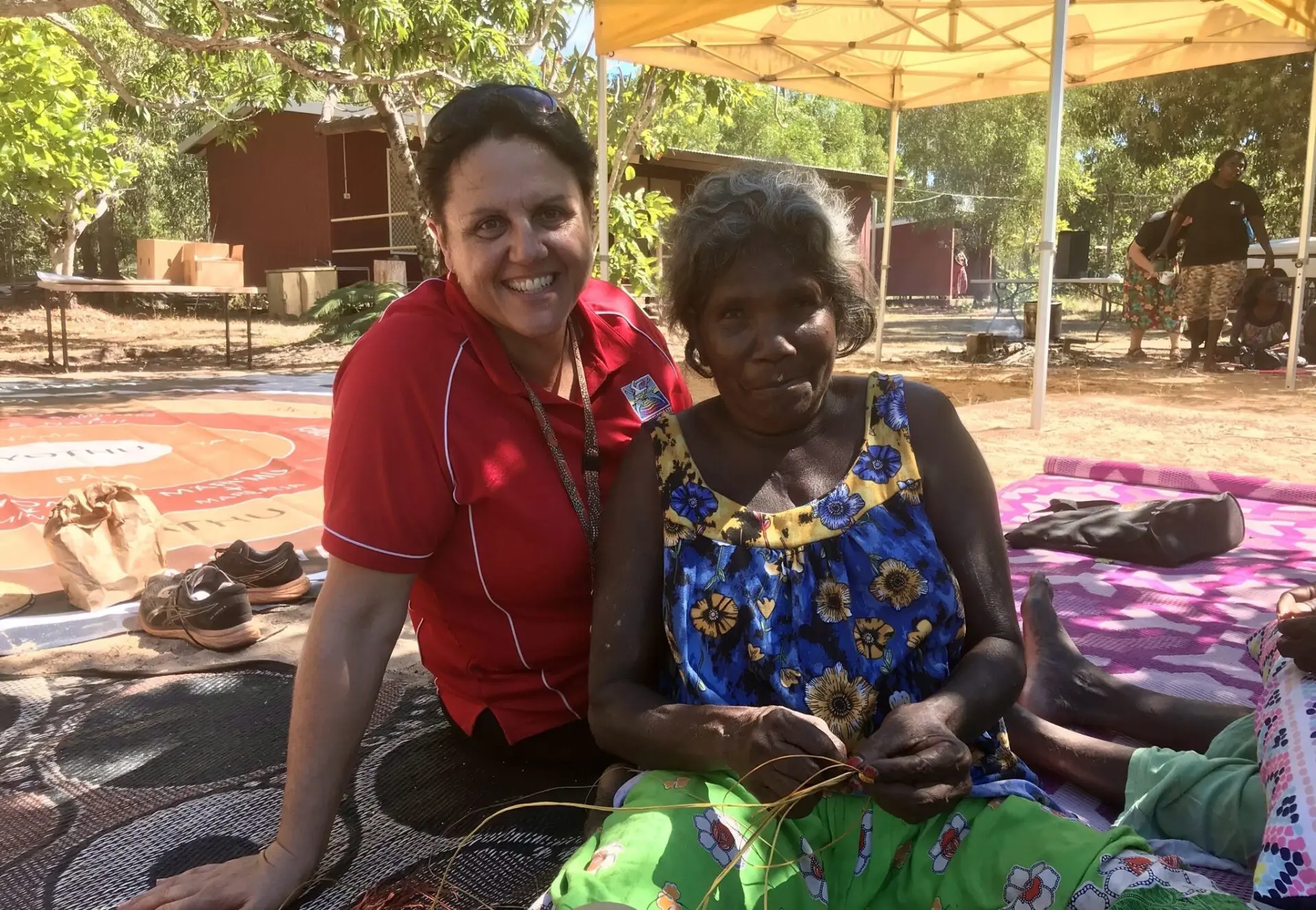 Kellie sat on a mat with a community member who is weaving.