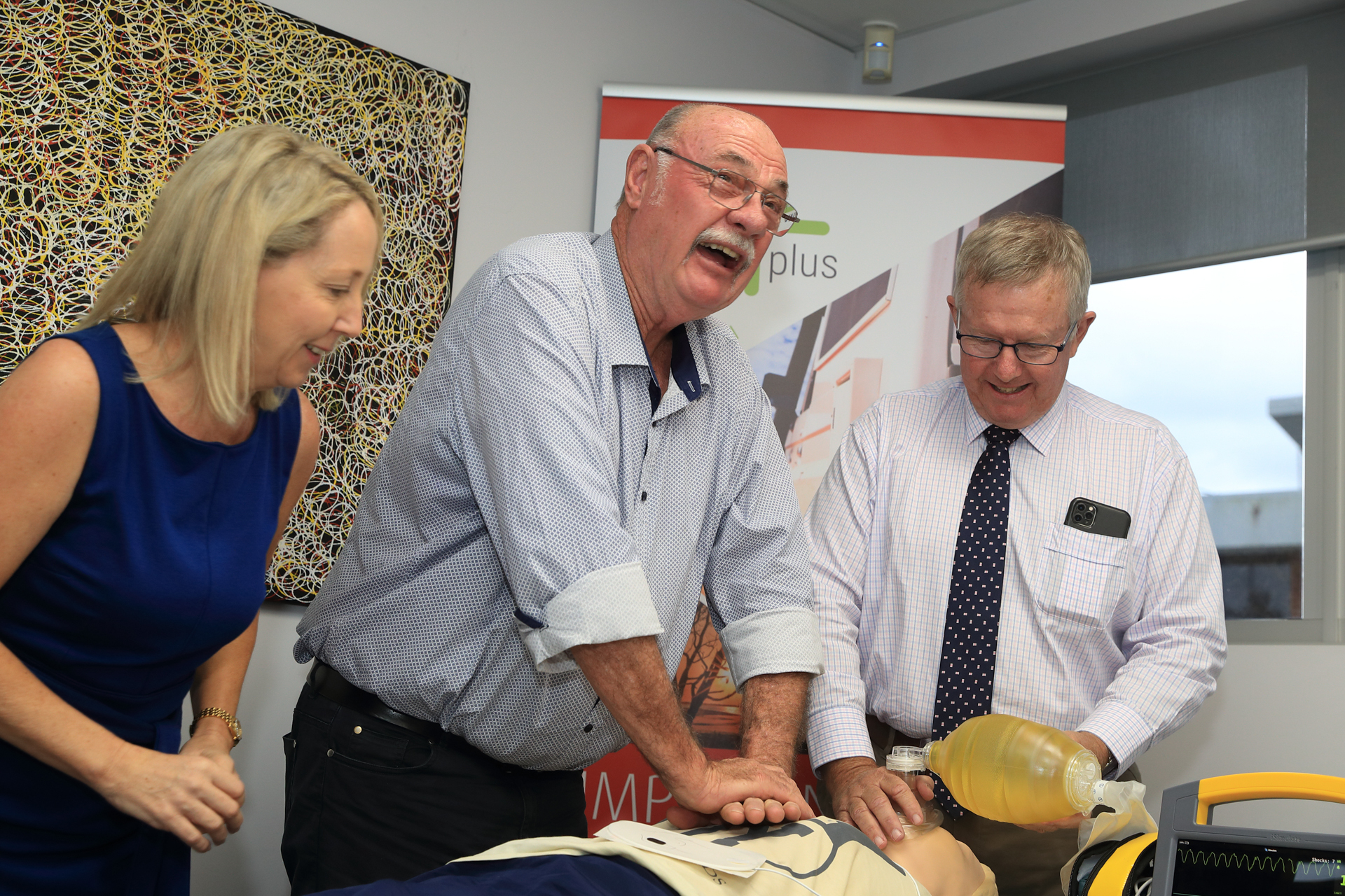 Man in shirt performs CPR on a mannequin. Another man in shirt holds air mask over mannequin's mouth. Woman in dress watches.