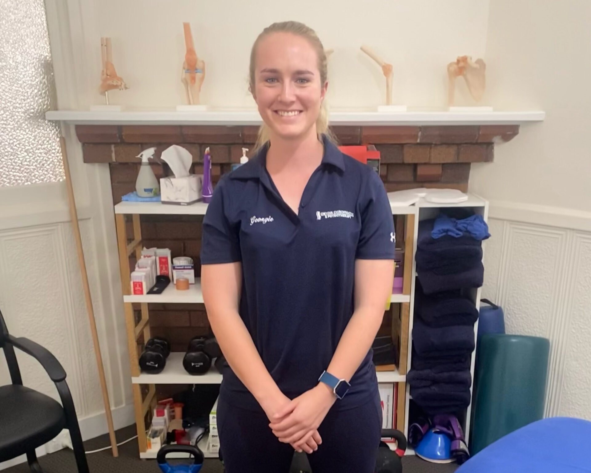 Georgina Haire. Georgina has blonde hair pulled back into a ponytail, and is stood in front of medical supplies.