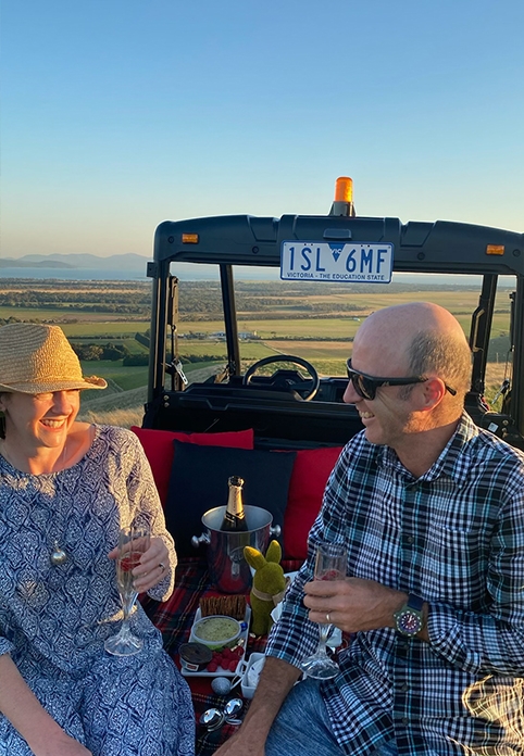 Two people sat on the back of a tractor, with a cheese platter and bottle of champagne