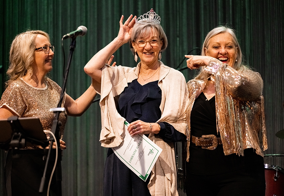 Lorraine Harry holding certificate and wearing tiara, with two women in sparkly tops smiling and pointing at her