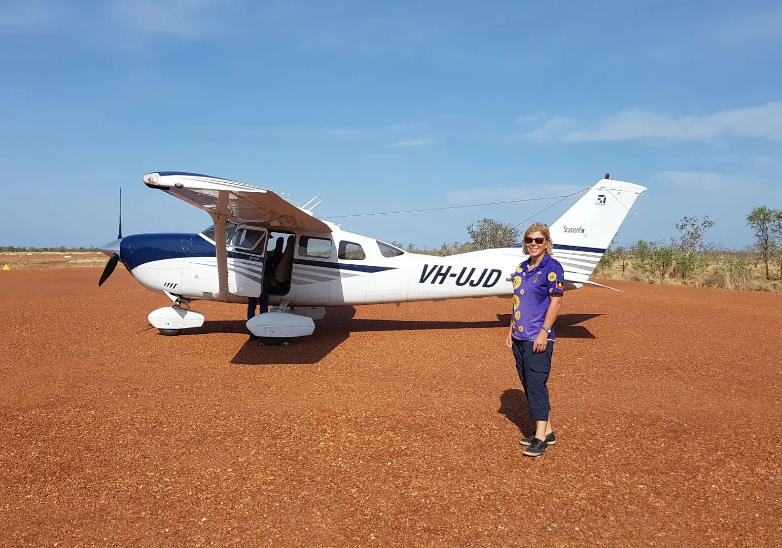 Woman standing in front of a small aircraft on red soil.