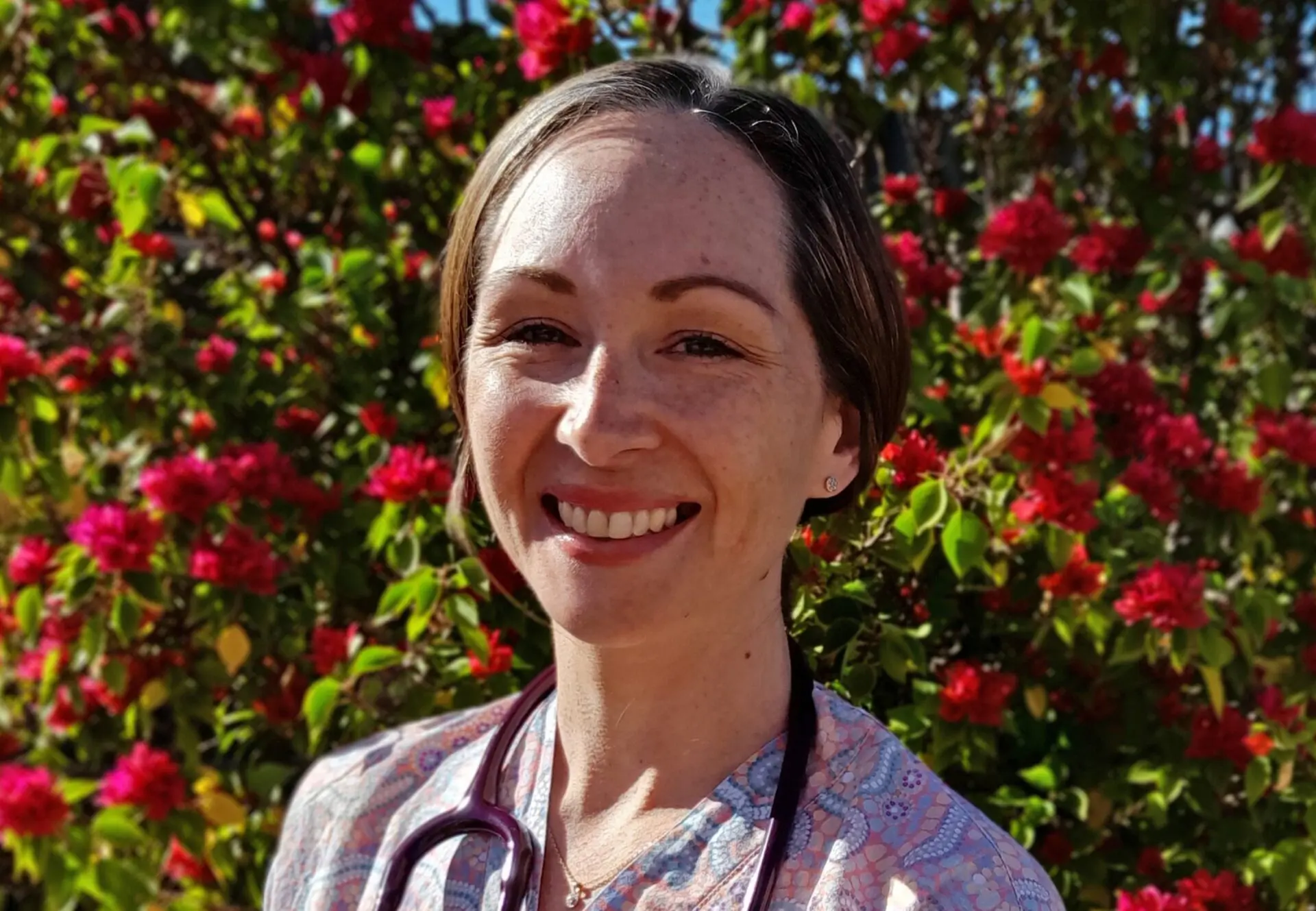 Jess standing in front of a bush of flowers, stethoscope around neck and hair pulled back