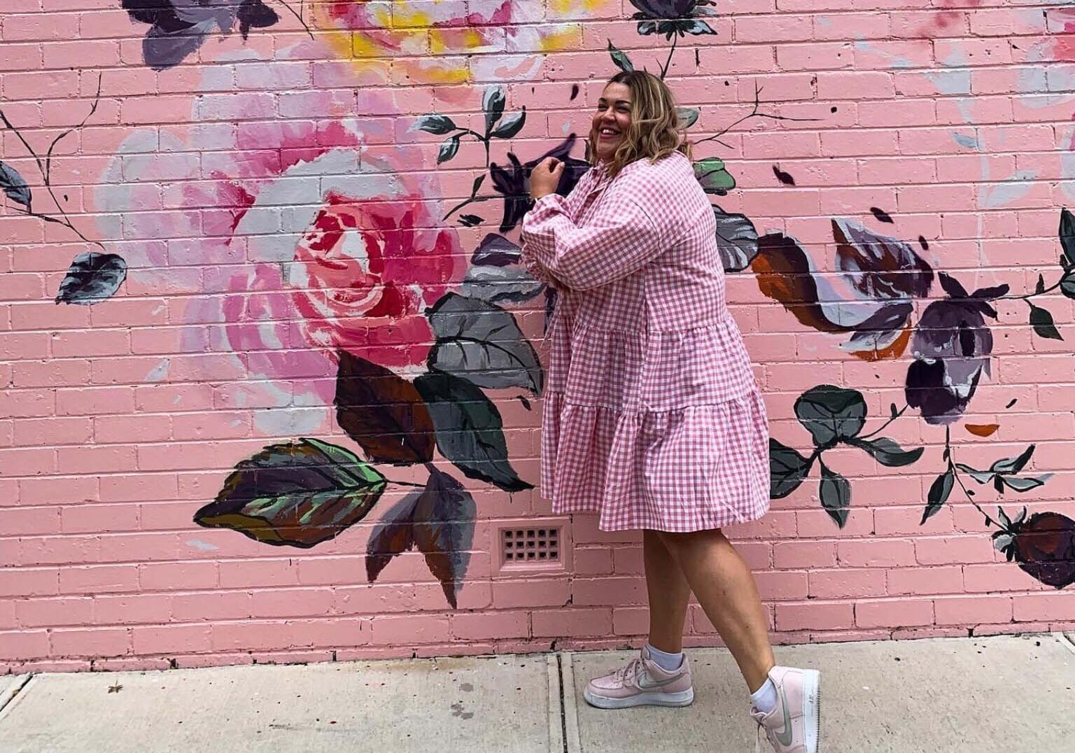 Woman in pink gingham dress standing in from of floral mural.
