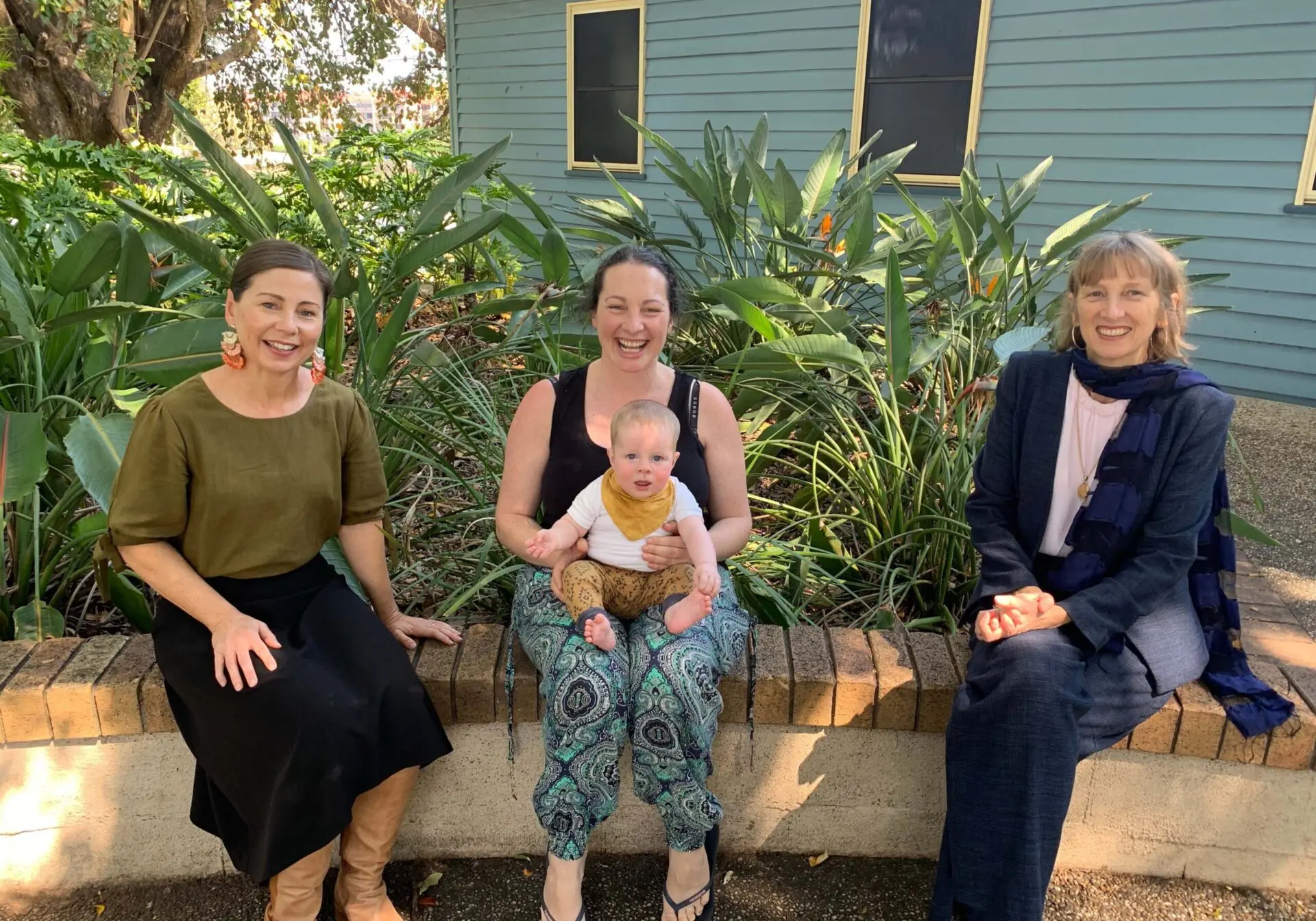 Three women sat on a garden bench, one holding a baby in her lap.