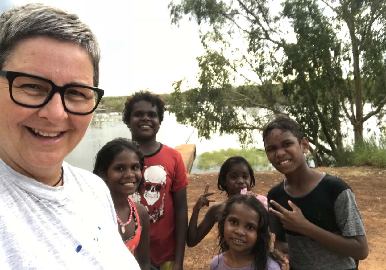 Woman taking a photo with children from community, smiling and putting up peace signs with their hands.