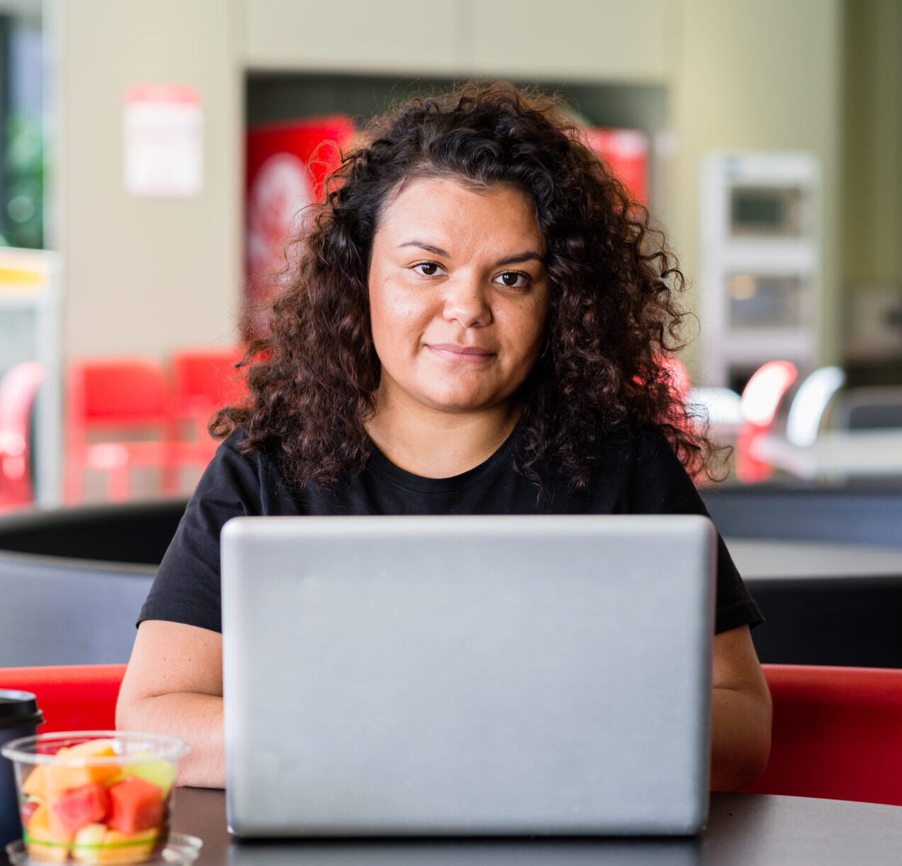 Young woman working on her laptop. The woman has dark curly hair and is wearing a black tshirt.